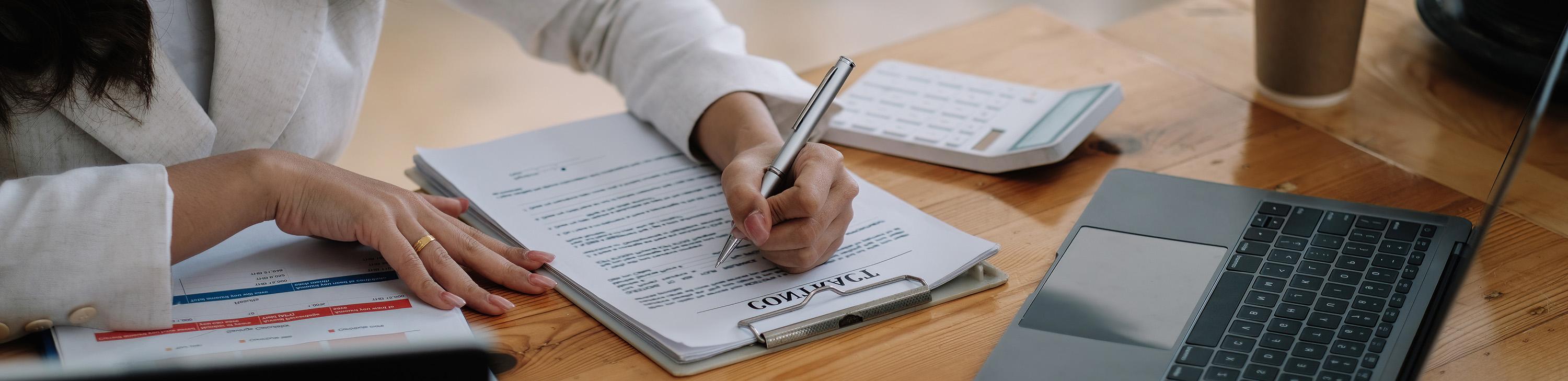 Hand signing contract with pen near laptop.